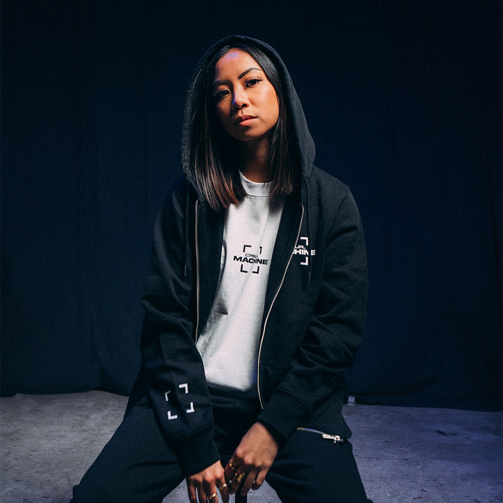 Mannequin femme assise sur une chaise, portant le Hoodie Basics 2.0 noir zippé avec la capuche relevée, coupe oversize et détails de marquage sérigraphié