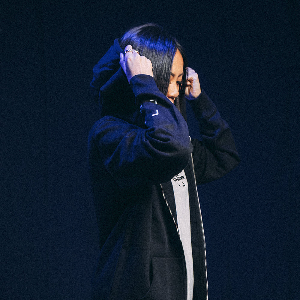 Mannequin femme de profil remettant la capuche du Hoodie Basics 2.0 noir zippé, marquage sérigraphié côté cœur et coupe unisexe oversize
