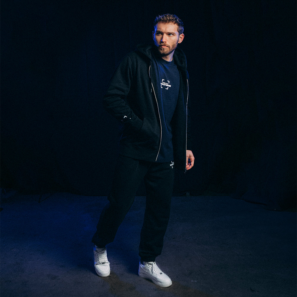 Homme portant le Jogging Basics 2.0 noir avec une veste assortie, coupe unisexe en coton organique, sérigraphie discrète sur la jambe gauche